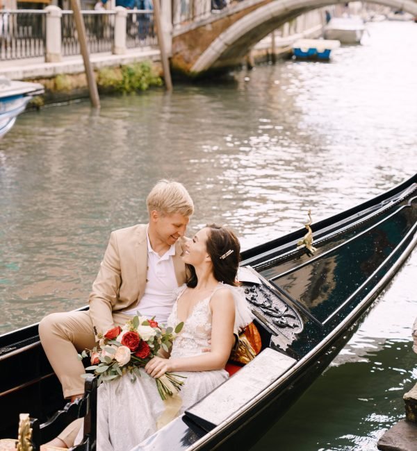 The bride and groom ride in a classic wooden gondola along a narrow Venetian canal. A close-up of cuddling newlyweds.
