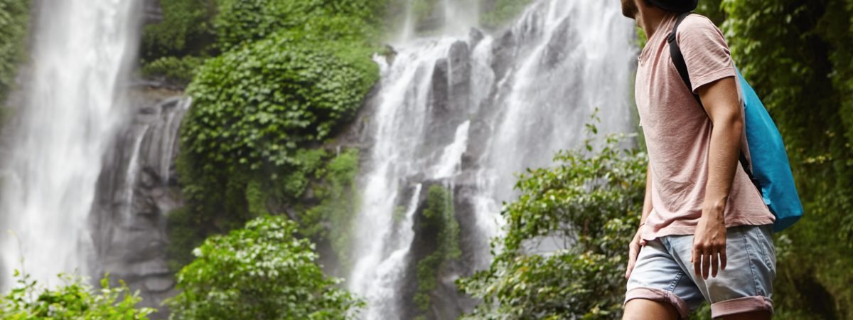 People, wildlife and adventure concept. Fashionable young adventurer with backpack contemplating waterfall landscape. Male tourist enjoying beautiful nature around him during his journey in rainforest