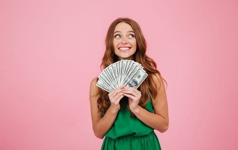 Portrait of a beautiful happy woman with long hair holding bunch of money banknotes and looking away at copy space isolated over pink background